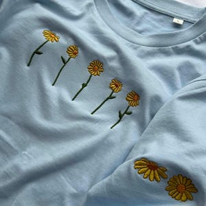 Peut inclure: T-shirt bleu clair avec des marguerites jaunes brod&eacute;es. Les marguerites sont dispos&eacute;es en rang&eacute;e sur la poitrine et sur la manche. Les fleurs ont des p&eacute;tales jaunes et des tiges vertes.