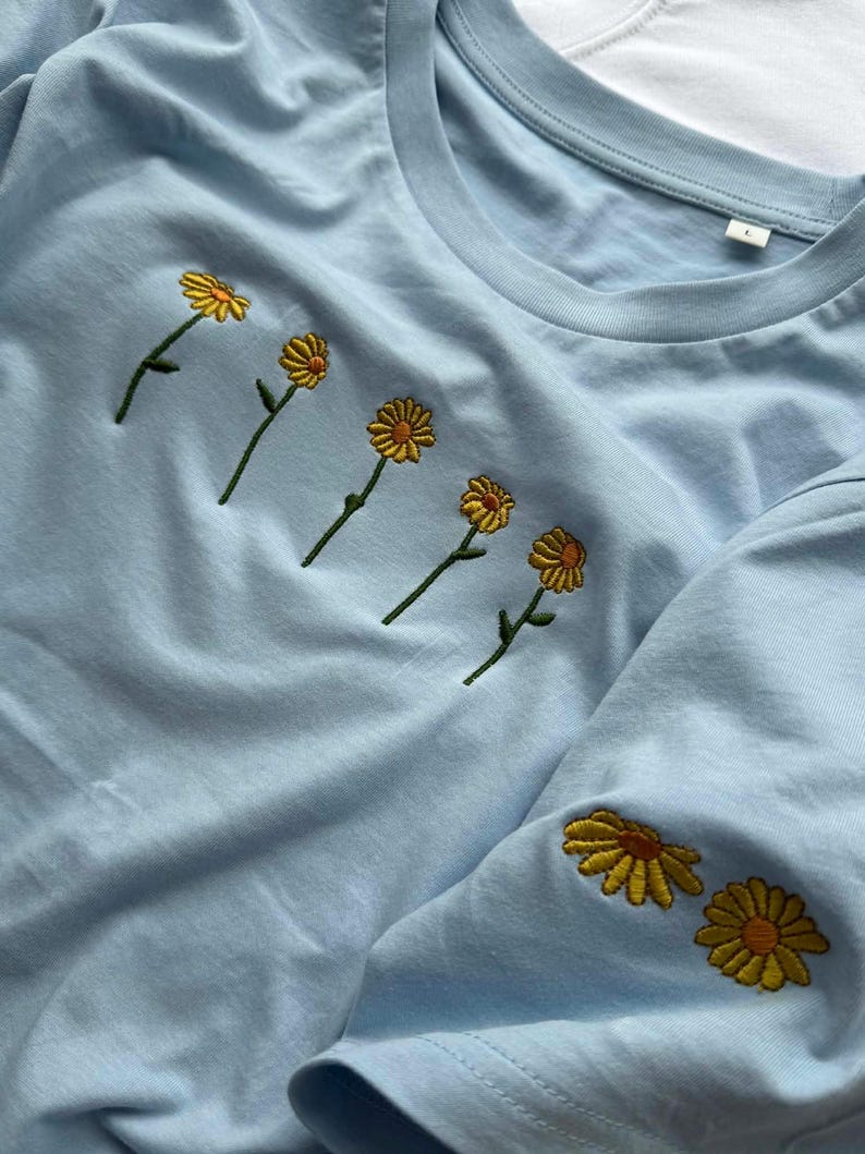 Peut inclure: T-shirt bleu clair avec des marguerites jaunes brod&eacute;es. Les marguerites sont dispos&eacute;es en rang&eacute;e sur la poitrine et sur la manche. Les fleurs ont des p&eacute;tales jaunes et des tiges vertes.