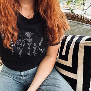 May include: Black t-shirt with a white floral design. The shirt has a crew neck and short sleeves. The person in the image is wearing blue jeans. A black and white geometric patterned cushion is in the background.