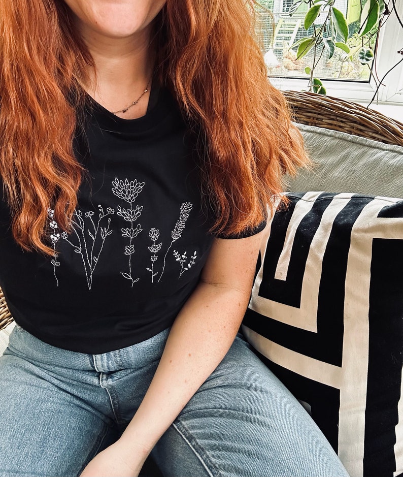 May include: Black t-shirt with a white floral design. The shirt has a crew neck and short sleeves. The person in the image is wearing blue jeans. A black and white geometric patterned cushion is in the background.