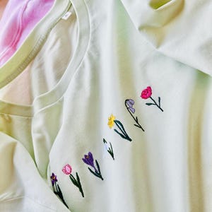 Peut inclure: Un t-shirt vert clair avec des fleurs brodées de différentes couleurs, dont le rose, le violet, le jaune et le blanc.