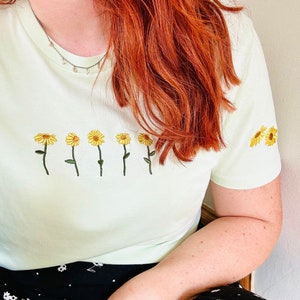 Peut inclure: T-shirt vert clair orn&eacute; de broderies de tournesols jaunes. Les fleurs sont dispos&eacute;es en rang&eacute;e sur le devant et sur la manche. Le t-shirt est de style d&eacute;contract&eacute;.