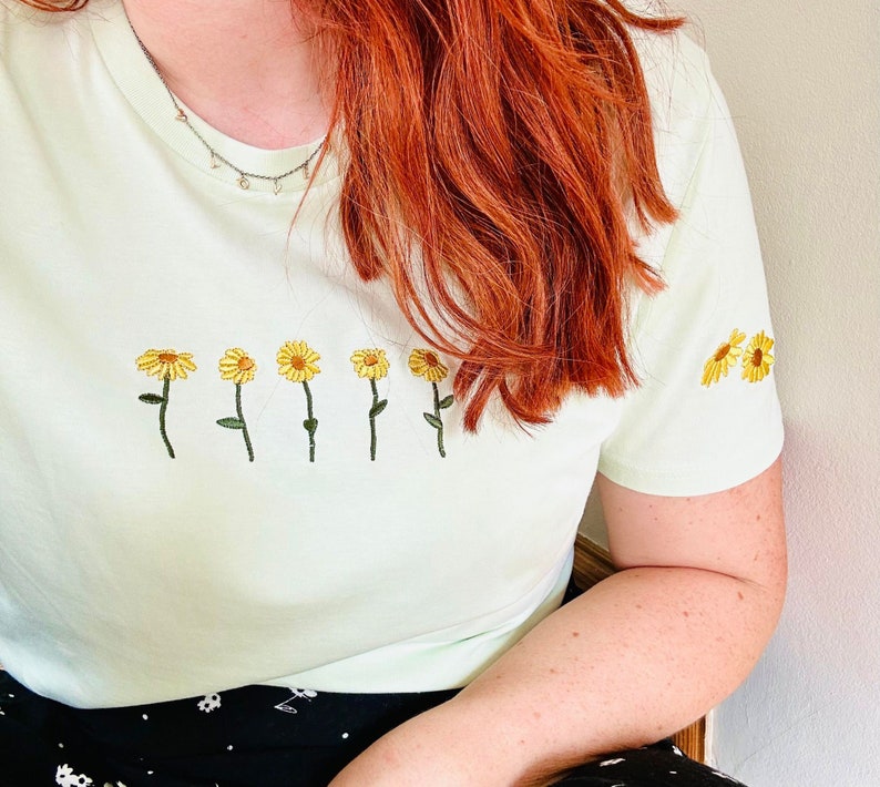 Peut inclure: T-shirt vert clair orn&eacute; de broderies de tournesols jaunes. Les fleurs sont dispos&eacute;es en rang&eacute;e sur le devant et sur la manche. Le t-shirt est de style d&eacute;contract&eacute;.