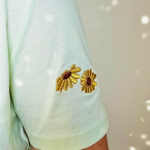 Peut inclure: Un t-shirt vert clair &agrave; manches courtes avec deux tournesols brod&eacute;s sur la manche. Les tournesols ont des p&eacute;tales jaunes et des centres bruns. Le t-shirt est fait d'un tissu doux et de couleur claire.