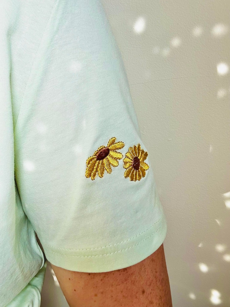 Peut inclure: Un t-shirt vert clair &agrave; manches courtes avec deux tournesols brod&eacute;s sur la manche. Les tournesols ont des p&eacute;tales jaunes et des centres bruns. Le t-shirt est fait d'un tissu doux et de couleur claire.
