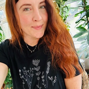 May include: Black t-shirt with a white floral embroidery design. The design features various flowers and foliage. The shirt is made of a soft material and is suitable for casual wear. The person in the image is wearing a gold necklace.