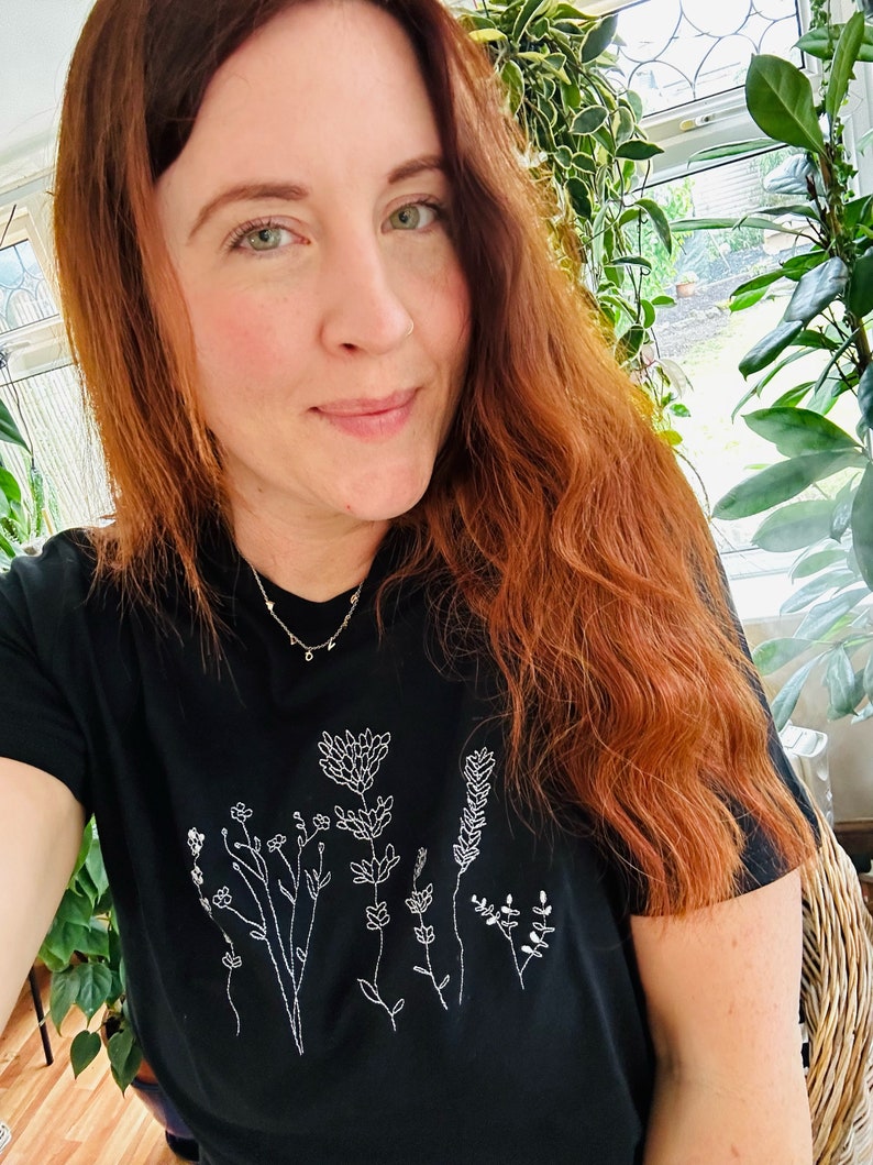 May include: Black t-shirt with a white floral embroidery design. The design features various flowers and foliage. The shirt is made of a soft material and is suitable for casual wear. The person in the image is wearing a gold necklace.