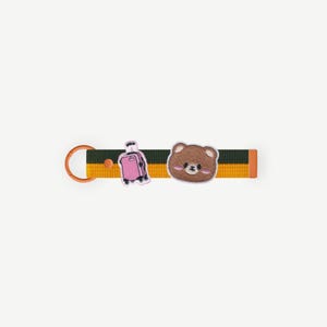Pode incluir: Um porta-chaves com uma cara de urso castanho e um berloque de mala rosa. A correia é listrada em verde, amarelo e laranja. O porta-chaves é cor de cobre.