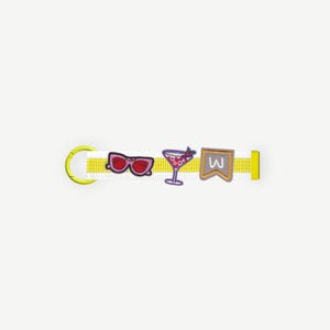 Puede incluir: Un collar para mascotas a rayas amarillas y blancas con un cierre de metal amarillo y una hebilla de ajuste de plástico amarillo. El collar presenta tres dijes: gafas de sol rosas, un cóctel y una pancarta con una "W".