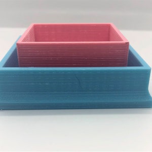 Puede incluir: Dos cajas cuadradas, una dentro de la otra. La caja exterior es de color azul claro y la caja interior es de color rosa claro.