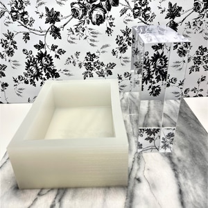 Puede incluir: Un molde de silicona blanco para hacer barras de jabón rectangulares. El molde está sobre una superficie de mármol junto a un bloque de vidrio transparente con un patrón floral.