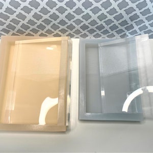 Puede incluir: Dos bandejas rectangulares de acrílico con tapas. La bandeja de la izquierda es de color beige claro y la bandeja de la derecha es de color gris claro.
