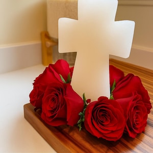 May include: A white cross-shaped candle with a wick, surrounded by vibrant red roses. The candle sits on a wooden base, creating a striking contrast. The image also includes the text "Cross-shaped Candle".