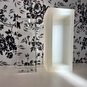 Può includere: Un blocco di cristallo rettangolare trasparente è accanto a uno stampo in schiuma rettangolare bianco. Il blocco di cristallo è in posizione verticale e riflette la luce. Lo sfondo presenta una carta da parati floreale in bianco e nero.