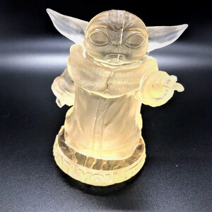 Puede incluir: Una figurita de pl&aacute;stico transparente de un personaje de la franquicia Star Wars, conocido como "The Child" o "Baby Yoda". La figurita est&aacute; de pie sobre una base con las palabras "What you see is what you get" inscritas en ella.