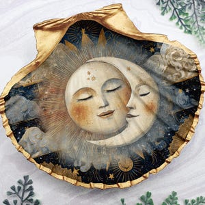 May include: A decorative seashell trinket dish with a celestial design. The interior features a sun and moon embracing, painted in soft colours against a starry night sky. The shell's edge is trimmed with gold.