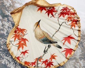 Könnte beinhalten: Dekorative Muschel mit goldfarbenem Rand, die einen gemalten Vogel auf einem Ast mit roten Ahornblättern zeigt. Der Vogel hat braune, blaue und weiße Federn. Der Hintergrund ist eine Landschaftsszene.