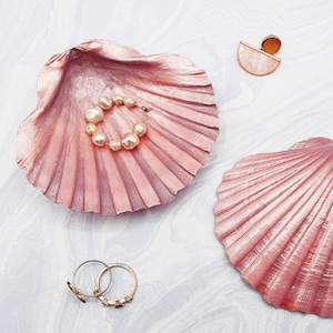 May include: Two pink seashell-shaped dishes, one with a pearl necklace and the other empty. The shells are made of a smooth, shiny material and have a delicate, feminine look.