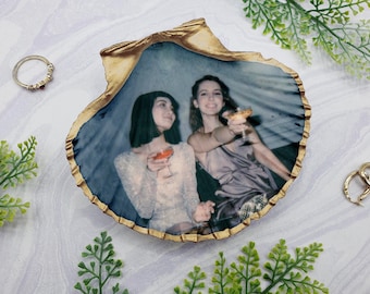 May include: Two women are pictured in a gold-rimmed seashell-shaped resin tray. The women are holding cocktails and smiling at the camera. The tray is a light blue color with a white and gold rim.