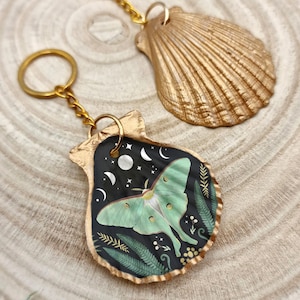 May include: A gold-colored seashell keychain with a black background featuring a green moth design, crescent moons, and stars. A second gold seashell pendant is visible in the background. The keychain has a gold chain and ring.