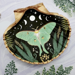 Puede incluir: Un plato de concha dorada con fondo negro y una polilla luna verde con marcas blancas. El plato está decorado con estrellas doradas, lunas y helechos.