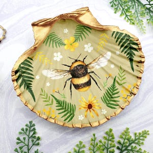 May include: A gold-rimmed seashell dish with a painted design of a bumblebee surrounded by green ferns and yellow flowers.