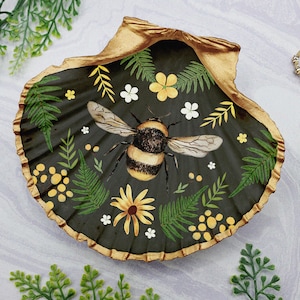 Puede incluir: Un plato de concha de mar negro y dorado con un diseño pintado de un abejorro, flores y helechos.