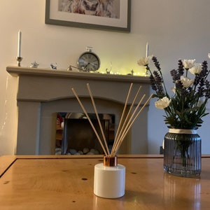May include: White ceramic diffuser bottle with a copper top and wooden sticks, sitting on a wooden table. A vase with white and purple flowers is in the background.