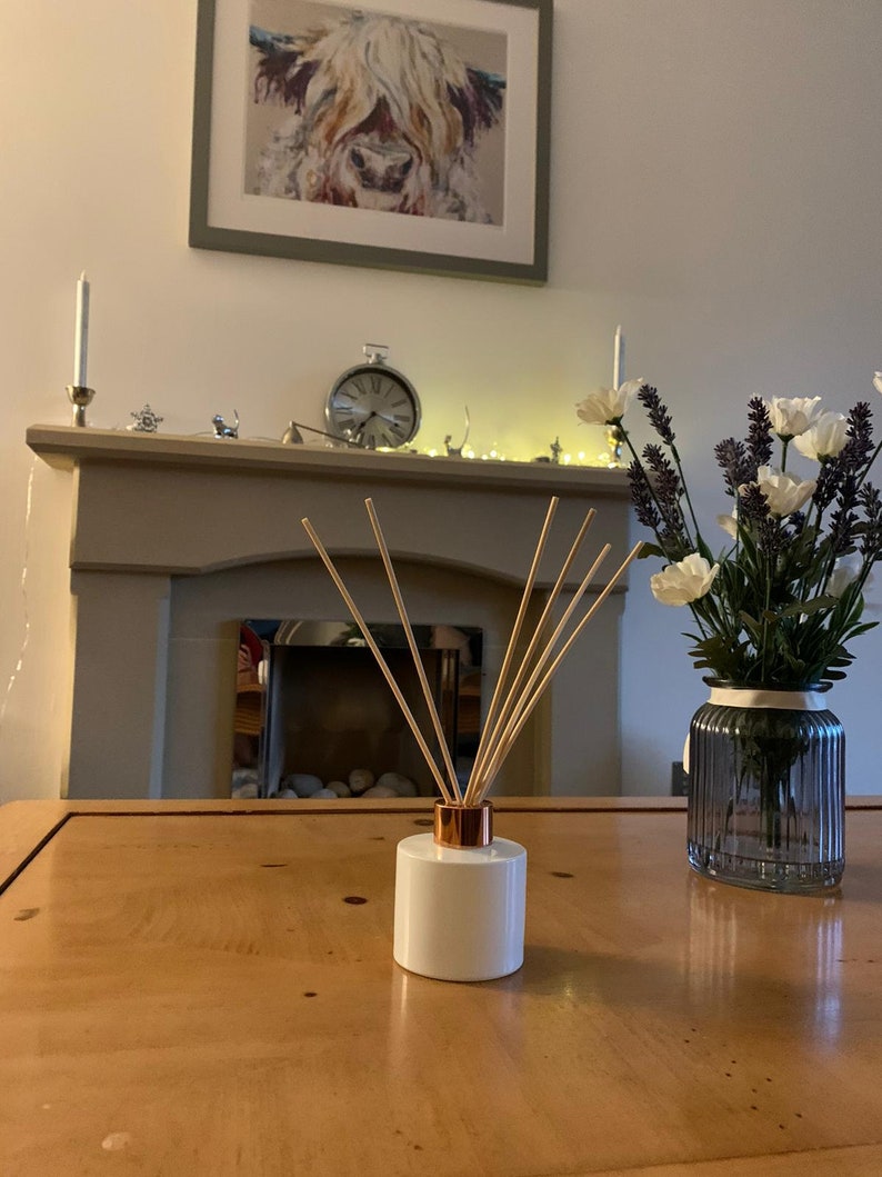 May include: White ceramic diffuser bottle with a copper top and wooden sticks, sitting on a wooden table. A vase with white and purple flowers is in the background.