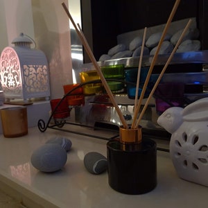 May include: A black diffuser bottle with wooden sticks and a gold top sits on a white surface with a white ceramic bunny, gray stones, and a candle holder with colorful glass votives.