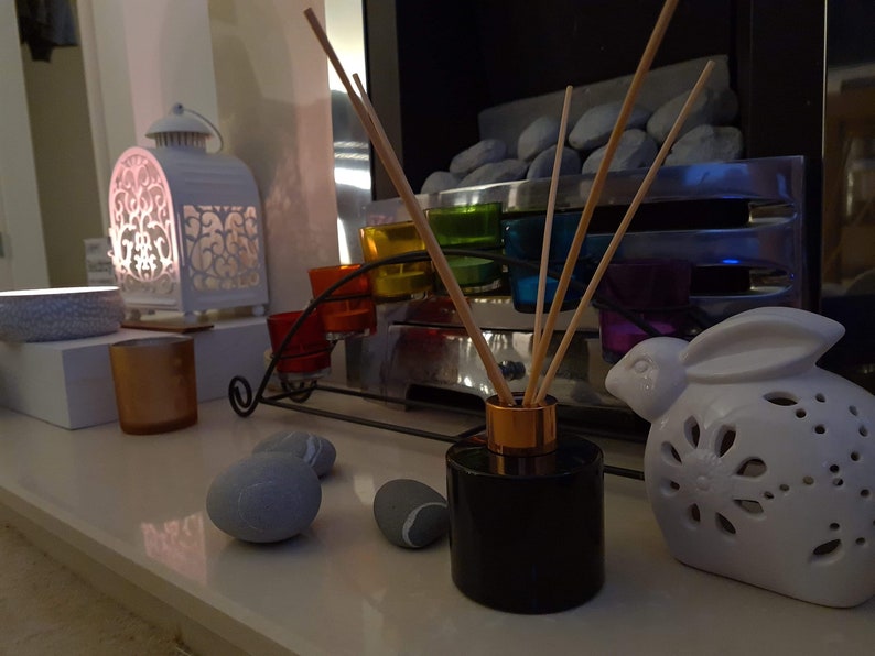 May include: A black diffuser bottle with wooden sticks and a gold top sits on a white surface with a white ceramic bunny, gray stones, and a candle holder with colorful glass votives.