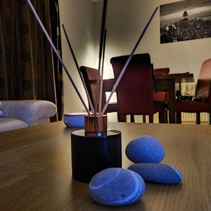May include: A black diffuser bottle with wooden sticks and a copper top sits on a wooden table with three gray stones. The diffuser is in the foreground of the image, with a white couch and a dining room set in the background.