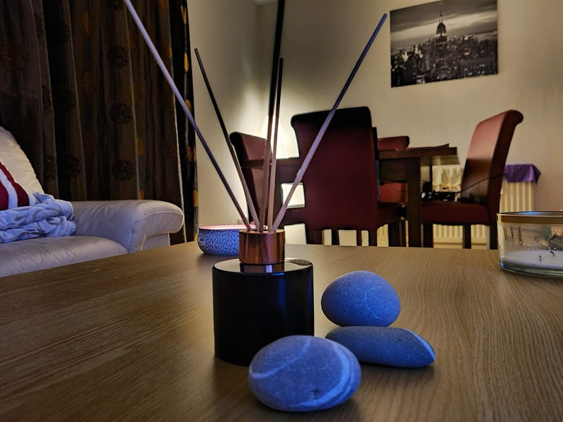 May include: A black diffuser bottle with wooden sticks and a copper top sits on a wooden table with three gray stones. The diffuser is in the foreground of the image, with a white couch and a dining room set in the background.