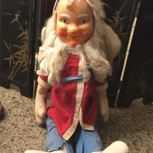 Puede incluir: Una muñeca vintage con una cara sonriente, cabello y barba blancos, y un abrigo rojo con ribete blanco. La muñeca lleva pantalones azules y zapatos blancos. La muñeca está sentada.