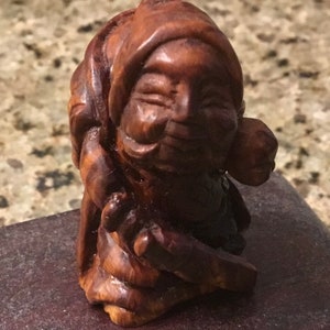 May include: A small, intricately carved wooden figurine depicting a smiling figure with a hooded head covering. The wood has a rich, dark brown color with visible grain, suggesting it is made from a hardwood. The figure's facial features are detailed.
