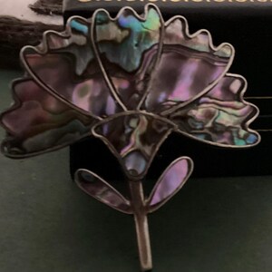 Pode incluir: Broche em forma de flor com pétalas de concha de abalone iridescente e uma moldura de metal prateado. O broche tem um caule e está sobre um fundo escuro. A concha de abalone exibe uma gama de cores, incluindo azuis, verdes e roxos.