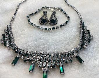 May include: A silver-toned jewelry set featuring a necklace, bracelet, and earrings. The necklace and bracelet are adorned with clear and emerald green rhinestones. The earrings have a teardrop design with a large green stone.