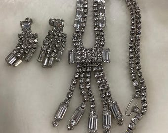 Op de afbeelding: Een zilverkleurige sieradenset met een ketting en bijpassende oorbellen. De ketting heeft een centraal rechthoekig ontwerp met bungelende kristallen accenten. De oorbellen zijn ook versierd met kristallen. De set wordt gepresenteerd op een witte ondergrond.