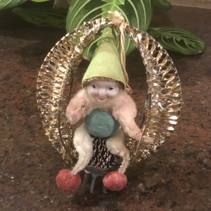 May include: A vintage Christmas ornament featuring a whimsical elf figure. The elf has a light green hat, a turquoise ball, and red pom-pom feet. It is suspended within a gold tinsel ring, creating a festive holiday decoration.