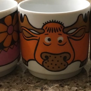 May include: White ceramic mugs with colorful cartoon animal designs. One mug features an orange moose, another has a pink and orange floral pattern, and a third has a yellow and orange design. The mugs are on a neutral surface.