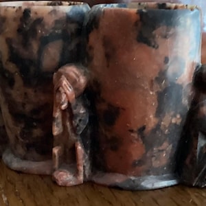 Puede incluir: Un portalápices de piedra tallada con dos compartimentos cilíndricos. El soporte es de color marrón rojizo con vetas negras y presenta tres pequeñas figuras de monos en varias poses. El portalápices está sobre una superficie de madera.