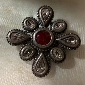 May include: A silver-toned brooch with a floral design. The center features a round, red gemstone, surrounded by clear, teardrop-shaped and oval-shaped stones. The brooch has a detailed, textured border.