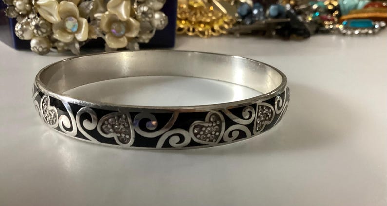 Op de afbeelding: Een zilverkleurige armband met een zwarte emaille inleg en een herhalend patroon van zilveren krullen en hartvormen. De hartmotieven zijn gevuld met kleine, heldere, reflecterende steentjes die het licht opvangen en een contrast vormen.