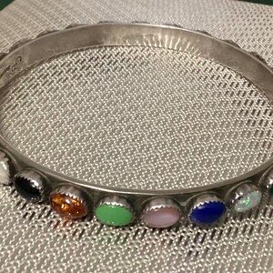 May include: A silver-toned bangle bracelet with a series of colorful gemstones. The gemstones are set in individual bezels and include a variety of colors such as red, green, blue, and white. The bracelet is resting on a textured surface.
