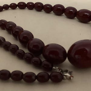 Op de afbeelding: Een ketting met gegradueerde, gladde, donkerbordeauxrode, ronde en ovale kralen. De kralen nemen in grootte toe naar het midden van de streng, die aan het uiteinde van de ketting is vastgezet met een kleine, metaalkleurige zilveren sluiting.