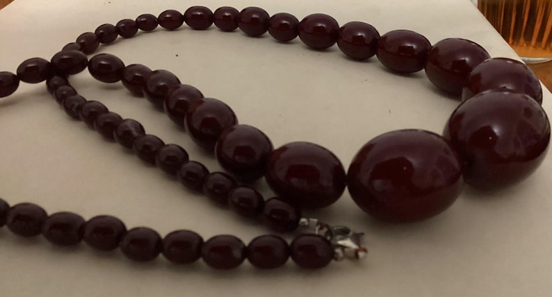 Op de afbeelding: Een ketting met gegradueerde, gladde, donkerbordeauxrode, ronde en ovale kralen. De kralen nemen in grootte toe naar het midden van de streng, die aan het uiteinde van de ketting is vastgezet met een kleine, metaalkleurige zilveren sluiting.