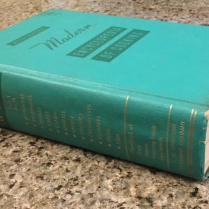 May include: A vintage turquoise cookbook titled "Modern Encyclopedia of Cooking." The book has gold lettering and a worn appearance, suggesting it's a well-loved kitchen reference. The book is resting on a speckled countertop.