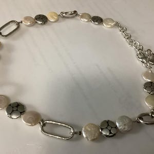May include: A necklace featuring alternating white coin-shaped pearls and round silver-toned metal discs with a floral-like embossed pattern. The design includes elongated oval silver-toned metal links and a silver-toned chain with a lobster clasp.