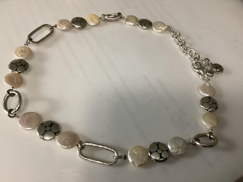 May include: A necklace featuring alternating white coin-shaped pearls and round silver-toned metal discs with a floral-like embossed pattern. The design includes elongated oval silver-toned metal links and a silver-toned chain with a lobster clasp.