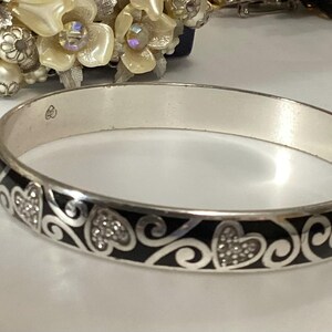 Op de afbeelding: Een zilverkleurige armband met een herhalend patroon van zwarte emaille krullen en hartvormen. De hartjes zijn gevuld met kleine, heldere, fonkelende steentjes. De binnenkant van de metalen band toont een klein gegraveerd keurmerk.